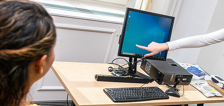 Trois personnes autour d’un bureau avec un ordinateur dans un environnement intérieur. Une personne est debout et pointe vers l’écran de l’ordinateur